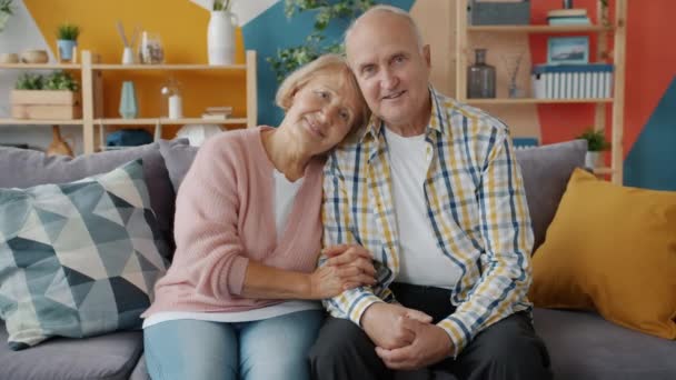 Портрет пары пожилых мужчин и женщин, улыбающихся, глядя в камеру дома на диване — стоковое видео