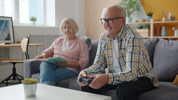 Медленное движение счастливой пожилой пары, играющей в видеоигры и читающей книгу дома — стоковое видео