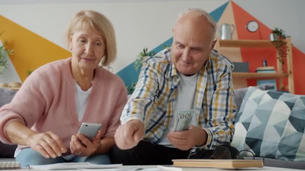 Старые муж и жена считают деньги, чтобы оплатить счета с помощью смартфона, а затем обнимаются — стоковое видео