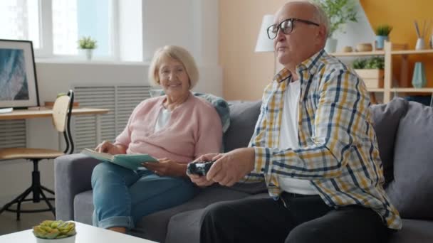 Mulher lendo livro enquanto homem idoso jogando videogame em casa no sofá — Vídeo de Stock