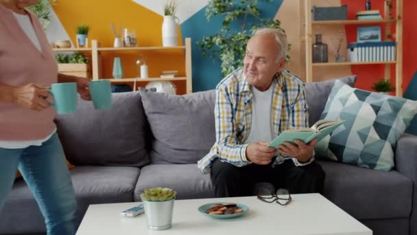 Zorgzame vrouw brengen thee en koekjes naar man lezen boek genieten van snack thuis — Stockvideo