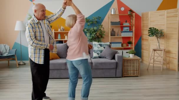 Cámara lenta de alegre pareja de ancianos bailando en casa tomados de la mano hablando sonriendo — Vídeos de Stock