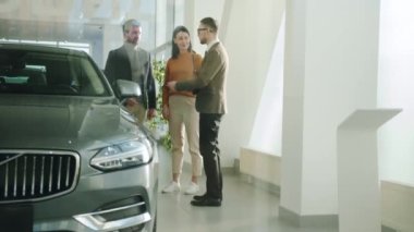 Girl and guy talking to salesman in car dealership choosing automobile together
