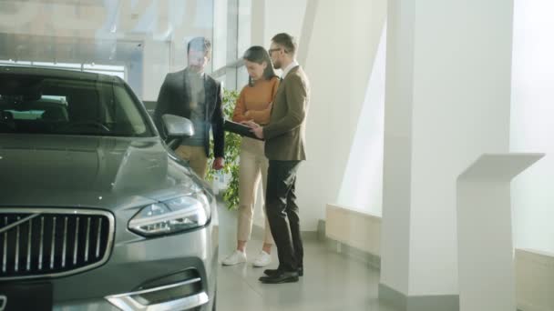 Happy young family husband and wife choosing auto in car dealership talking to salesman — Stock video