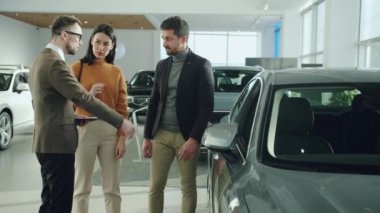 Confident salesman talking to cheerful young couple in car showroom showing automobile