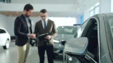 Business man talking to sales manager choosing car in contemporary showroom. Focus on vehicle mirror