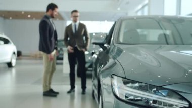 Handsome guy talking to sales agent in car dealership, focus on expensive automobile. Focus on car headlight