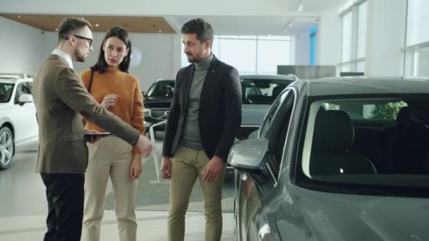 Confident salesman talking to cheerful young couple in car showroom showing automobile — Stock video