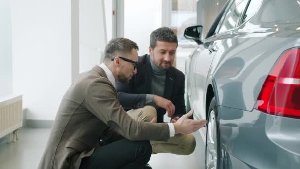 Медленное движение мужчин клиент и продавец обсуждают автомобиль в выставочном зале, рассматривая шины — стоковое видео