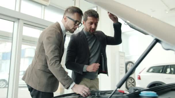 Менеджер по продажам и клиент проверяют двигатель под капотом, разговаривая в выставочном зале — стоковое видео