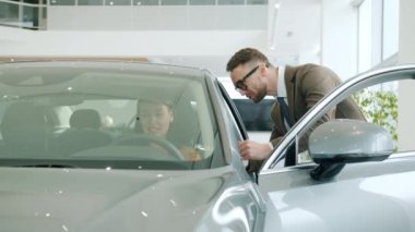 Attractive girl talking to salesman in motor dealership sitting in modern new car