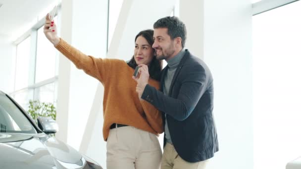 Gioioso uomo e donna che si scattano selfie con le chiavi della macchina in concessionaria utilizzando la fotocamera dello smartphone — Video Stock