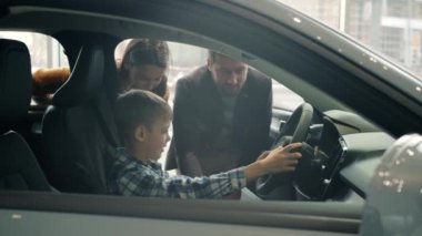 Joyful kid sitting in new car holding steering wheel while parents buying automobile