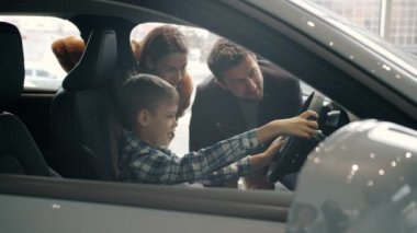 Happy child sitting in car in drivers seat holding steering wheel choosing auto with parents