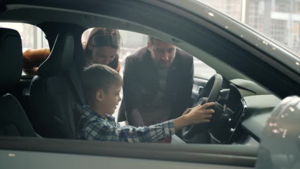 Alegre niño sentado en un coche nuevo sosteniendo el volante mientras los padres compran automóvil — Vídeos de Stock