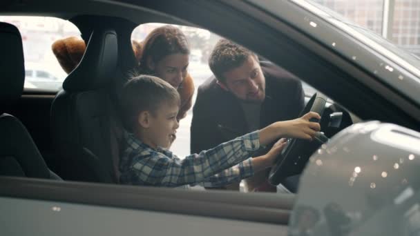 Happy child sitting in car in drivers seat holding steering wheel choosing auto with parents — Wideo stockowe