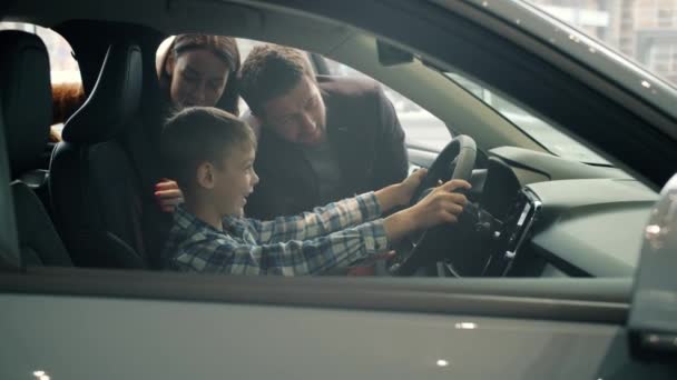 Семья с сыном покупает автомобиль в выставочном зале, ребенок веселится с рулем — стоковое видео