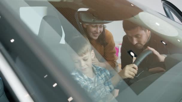 Hombre y mujer elegir el coche en la concesionaria, mientras que el hijo jugando con el volante — Vídeos de Stock