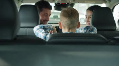 Joyful parents talking to little boy choosing car in showroom sitting inside automobile