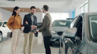 Handsome car buyer getting key fob from salesman shaking hands hugging girlfriend