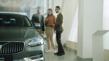 Young people buying car in motor showroom talking to salesperson looking at automobile