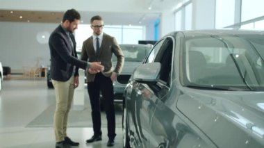 Cheerful businessman buying car in showroom talking to sales agent looking at auto. Focus on car mirror