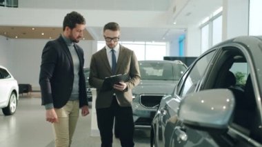 Confident sales manager talking to male customer in car dealership showing auto. Focus on vehicle mirror