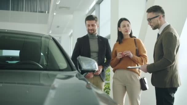 Счастливые мужчина и женщина разговаривают с автодилером в салоне обсуждая автомобили — стоковое видео