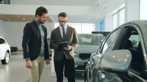 Gerente de ventas de confianza hablando con el cliente masculino en la concesionaria de automóviles que muestra auto. Enfoque en el espejo del vehículo — Vídeos de Stock