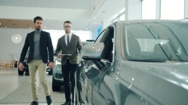 Friendly sales manager in car dealership talking to male customer showing automobile — Stockvideo