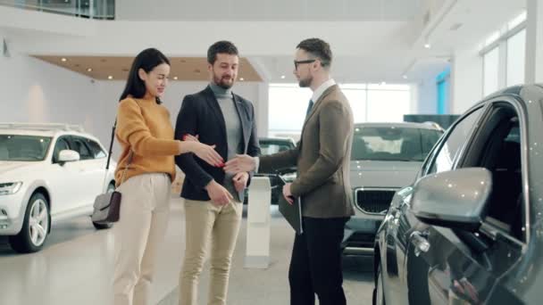 Slow motion of young couple buying car shaking hands with agent hugging taking keys — Stock video