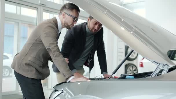 Cheerful men sales manager and client watching car engine under motor hood indoors — Αρχείο Βίντεο