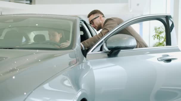 Cheerful young woman checking car interior in showroom talking to salesman — Αρχείο Βίντεο