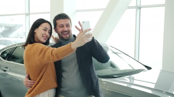 Bella giovane coppia prendendo selfie in auto showroom in posa con nuova auto sorridente — Video Stock