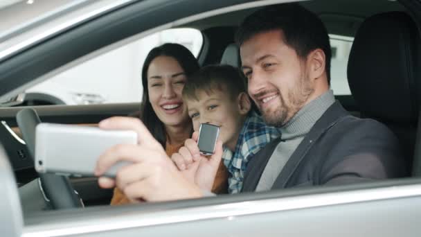 Happy young family with kid taking selfie in new auto using smartphone camera — Stock video
