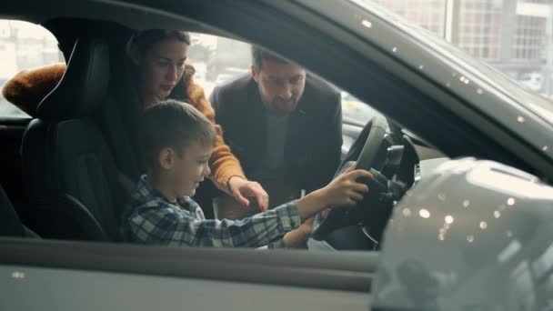 Очаровательный маленький ребенок за рулем автомобиля в автосалоне покупает автомобиль с родителями — стоковое видео