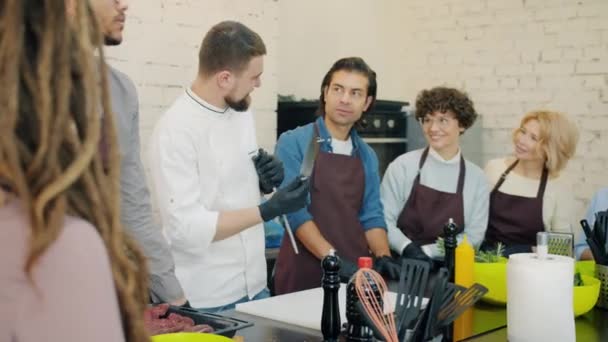 Chef alegre en uniforme afilando cuchillo y hablando con estudiantes de clase de cocina — Vídeos de Stock