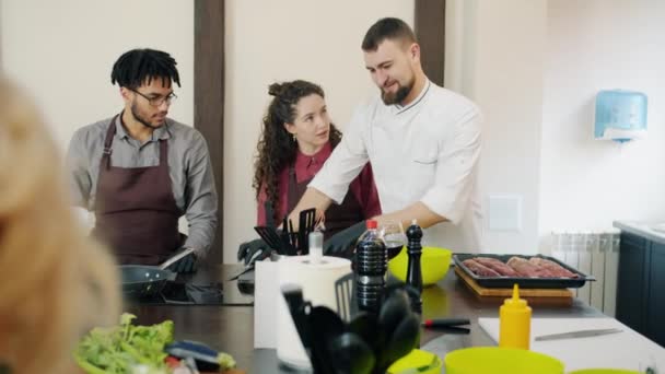 Разнообразная группа студентов кулинарии разговаривает с шеф-поваром, наблюдая, как он готовит еду — стоковое видео