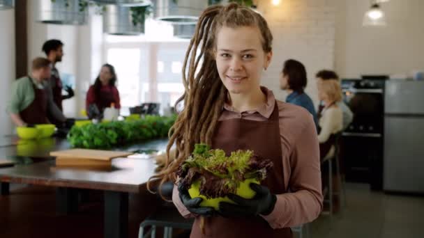 Πορτρέτο της ελκυστική νεαρή κοπέλα με dreadlocks χαμογελώντας στη σχολή μαγειρικής κρατώντας σαλάτα — Αρχείο Βίντεο