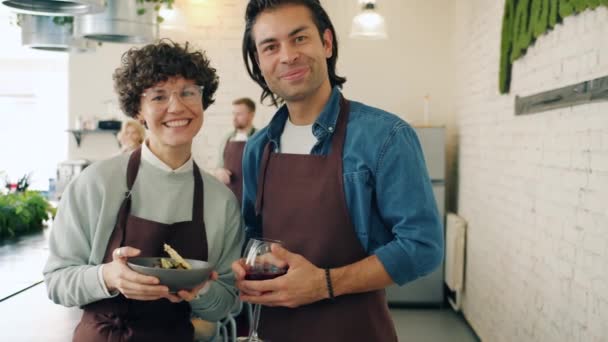 料理教室で食べ物や飲み物を持って笑顔エプロンで幸せな若いカップルの肖像画 — ストック動画