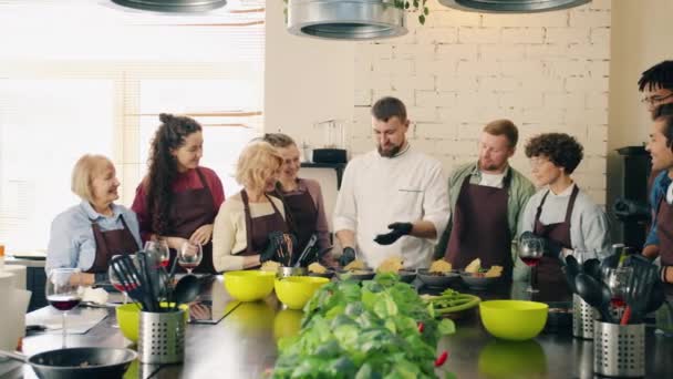 Группа парней и девушек хлопают в ладоши шеф-повару держа приготовленное блюдо на кухне — стоковое видео