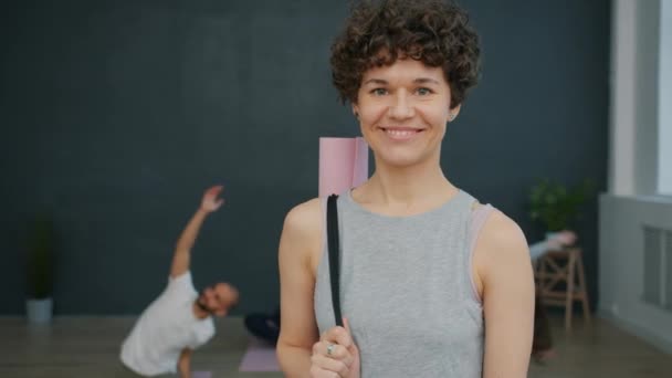 Портрет привлекательной девушки в спортивном наряде с ковриком для йоги, улыбающейся в студии — стоковое видео