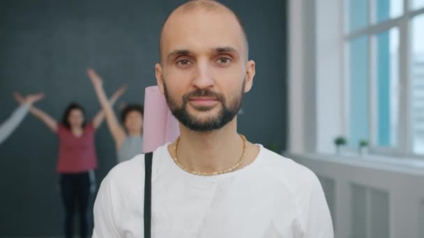 Портрет красивого молодого чоловіка з килимком йоги, який посміхається дивитися на камеру в студії — стокове відео