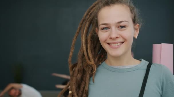 Portrait de jolie jeune femme avec dreadlocks tenant tapis de yoga dans la salle de gym souriant — Video