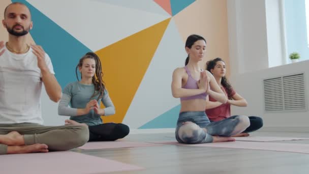 Chicas y chico disfrutando de la meditación durante la práctica de yoga en un acogedor estudio sentado en pose de loto — Vídeo de stock