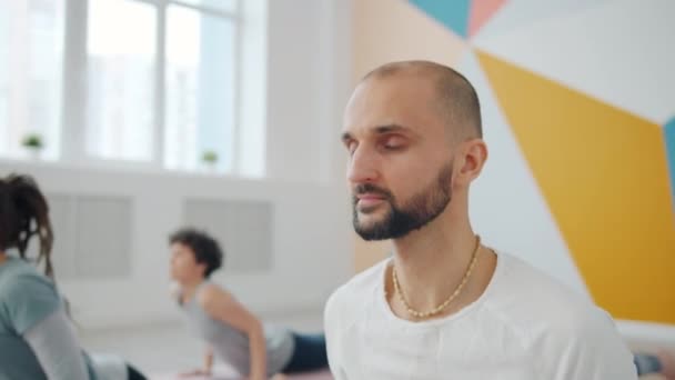 Бородатый парень занимается в помещении в классе йоги с молодыми женщинами, движущимися по коврику — стоковое видео