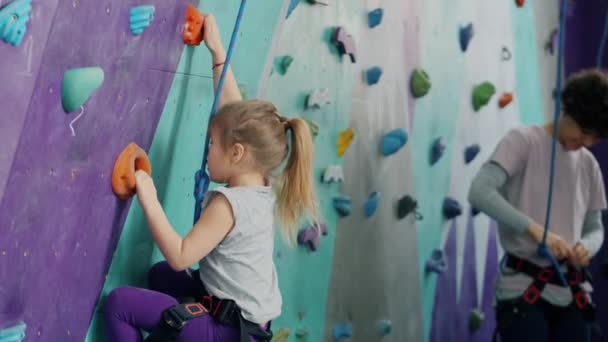 Klein kind klimmen kunstmatige muur in sportcentrum, vrouw met behulp van veiligheidsuitrusting op de achtergrond — Stockvideo