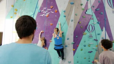 Çocuklu bir kadın spor salonunda tırmanırken insanlar ipleri tutarak eğleniyor.