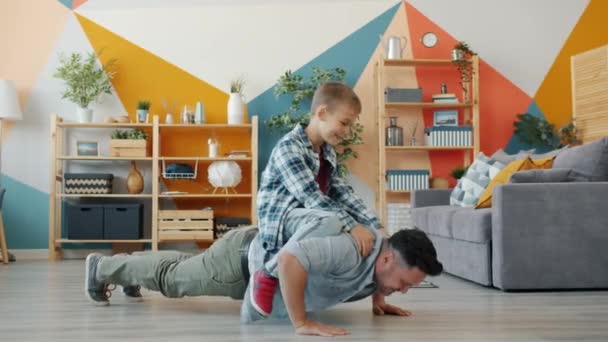 Fuerte padre hombre haciendo flexiones mientras hijo sentado en su espalda sonriendo en casa — Vídeos de Stock