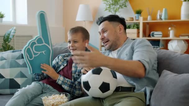 Fãs de futebol pai e criança assistindo jogo na TV em casa segurando mão espuma e bola — Vídeo de Stock